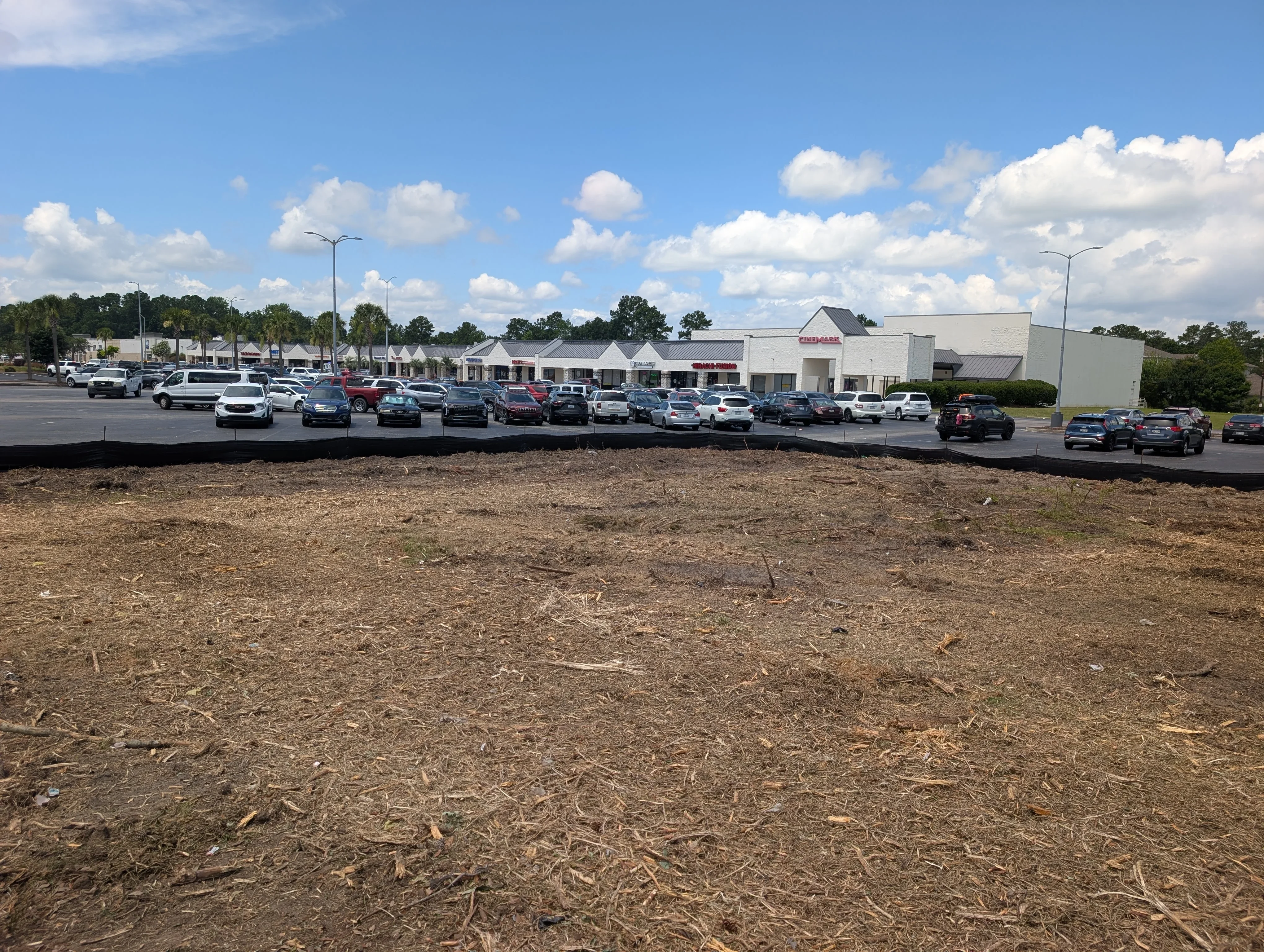 Graded dirt parking lot with compacted base, retail buildings and parked cars in background