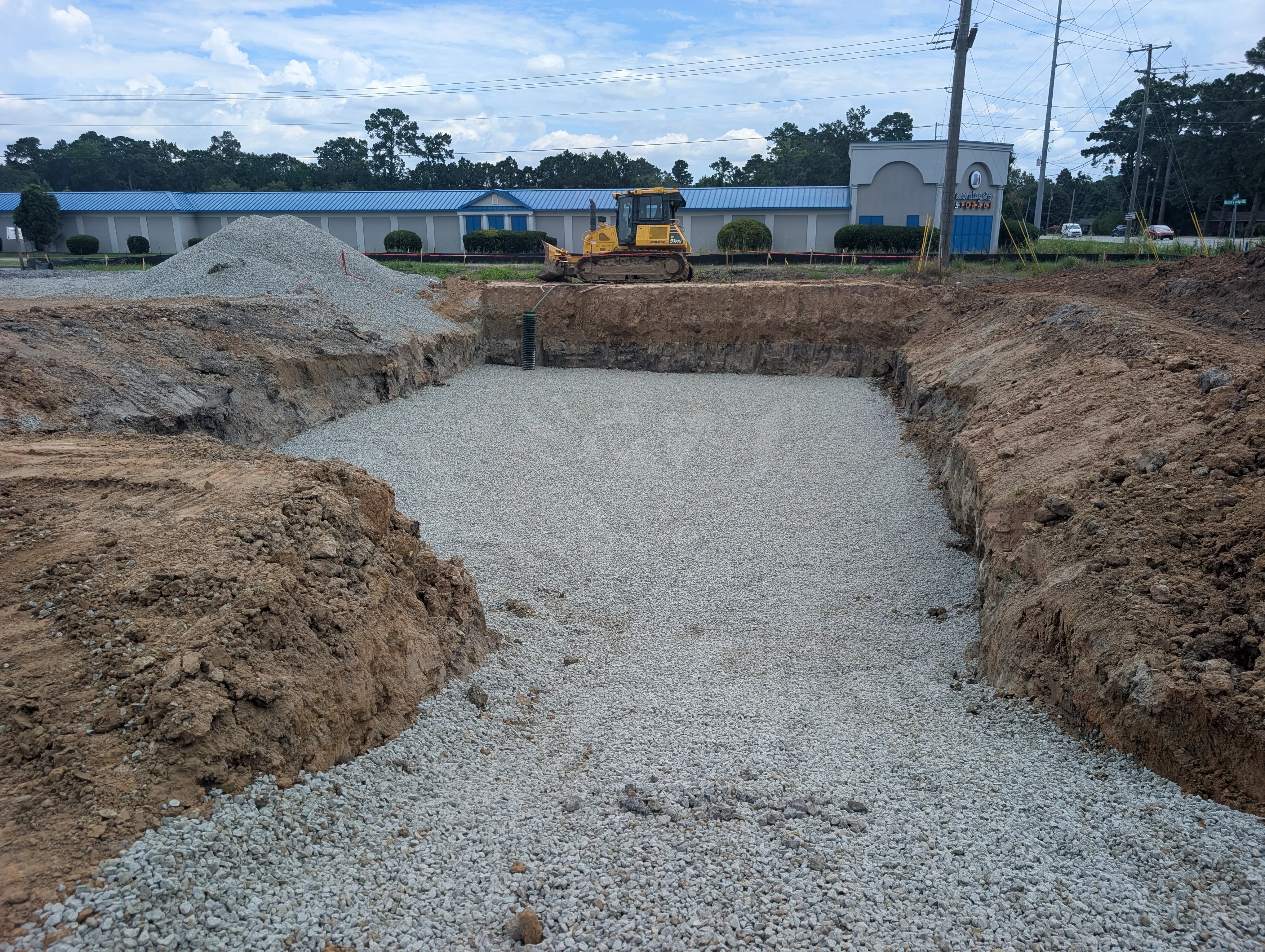Excavated pond with gravel base, sloped dirt walls, and drainage pipe installation