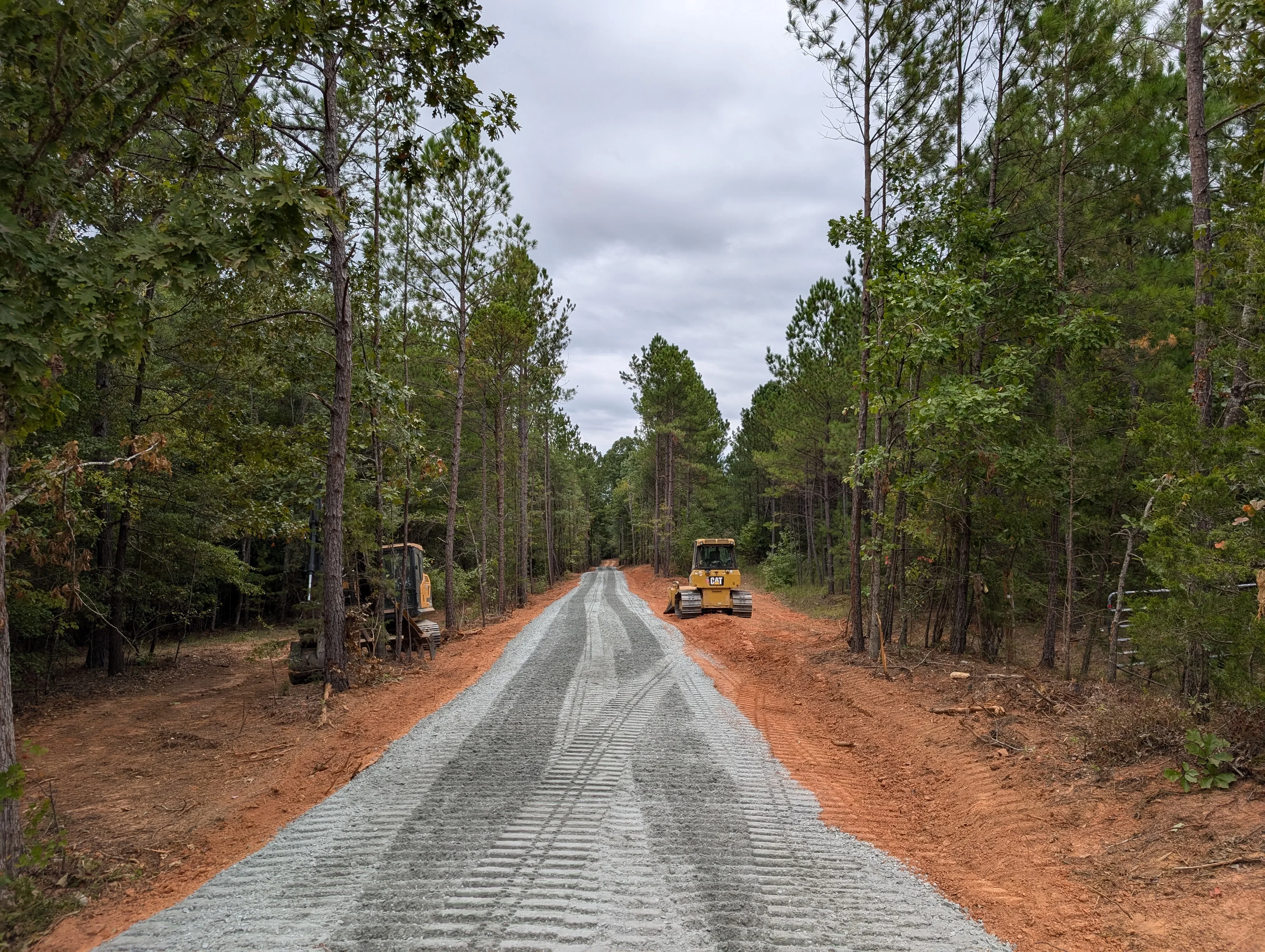 Graded forest road with crushed stone base, red clay shoulders, and heavy equipment in wooded area
