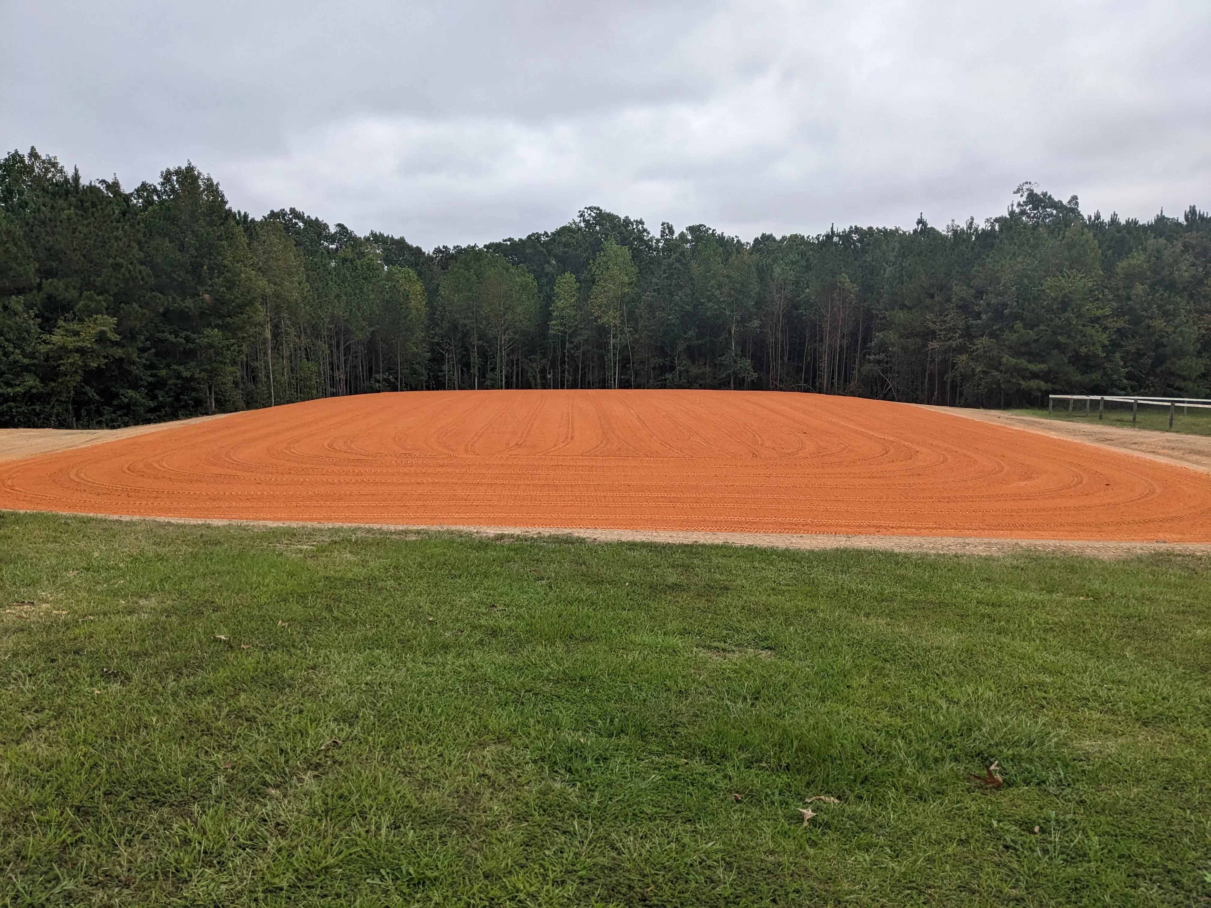 Freshly graded clay site with uniform compaction patterns and tree line backdrop