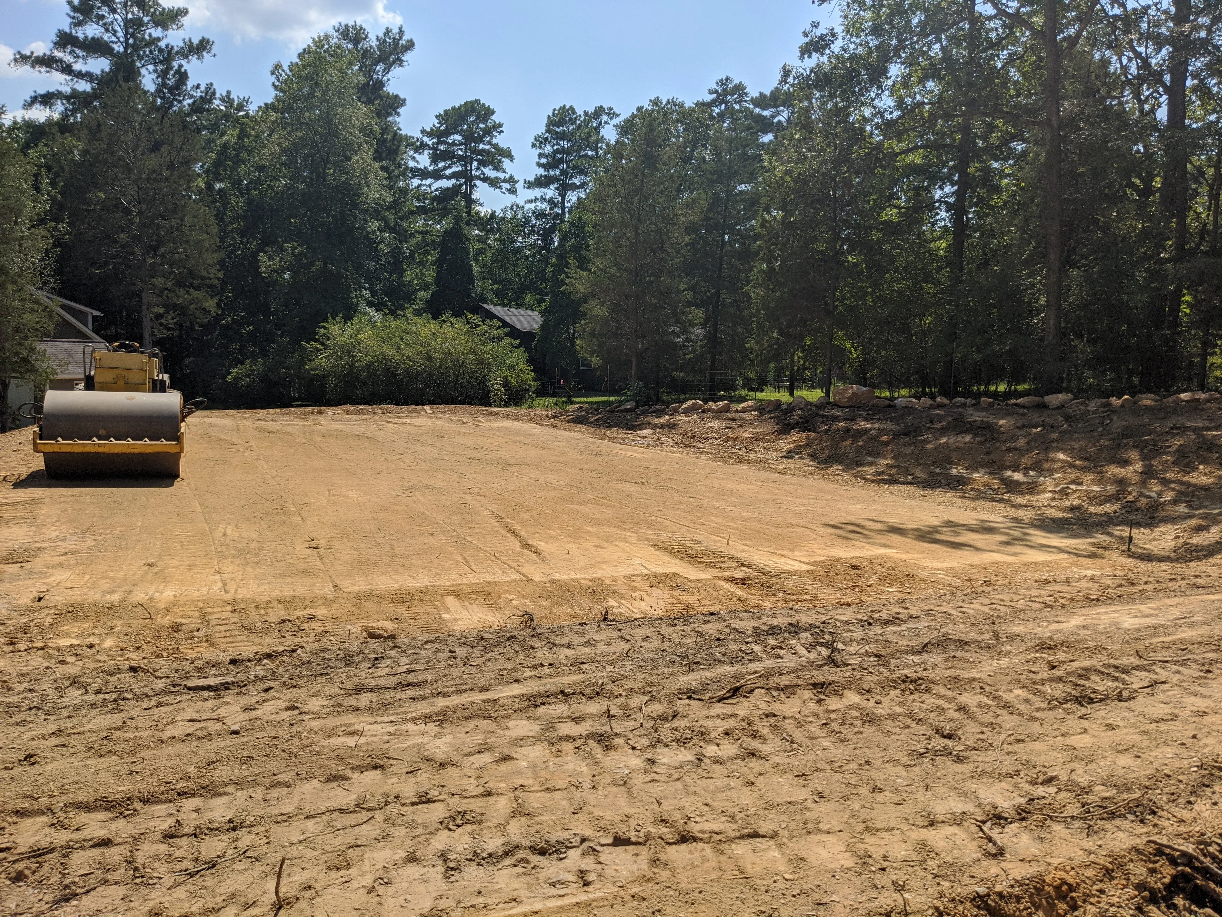 Vibratory roller compacting graded dirt driveway with cleared site and tree line in background