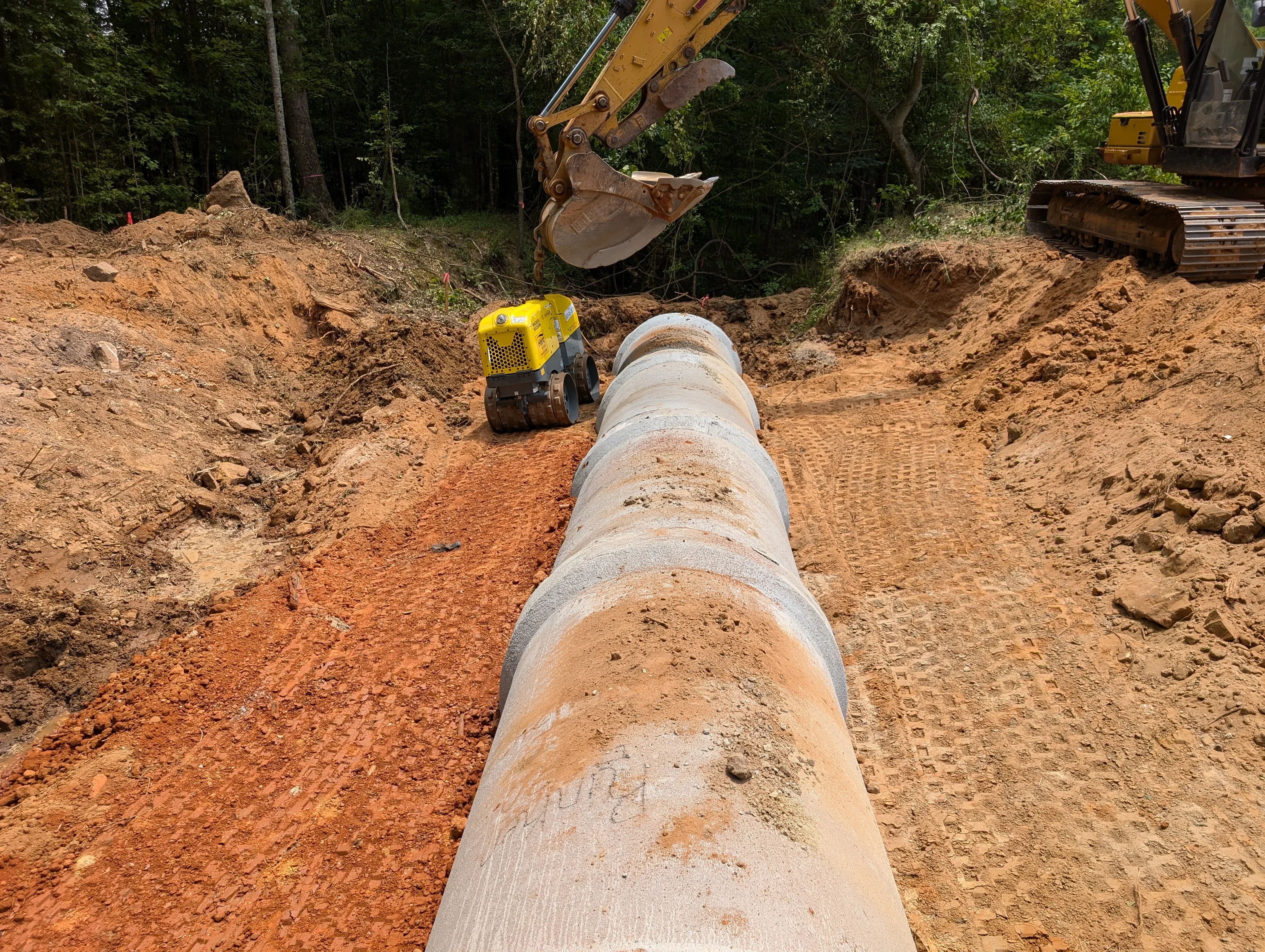 Concrete pipe laid in excavated trench with compactor and excavator on wooded site