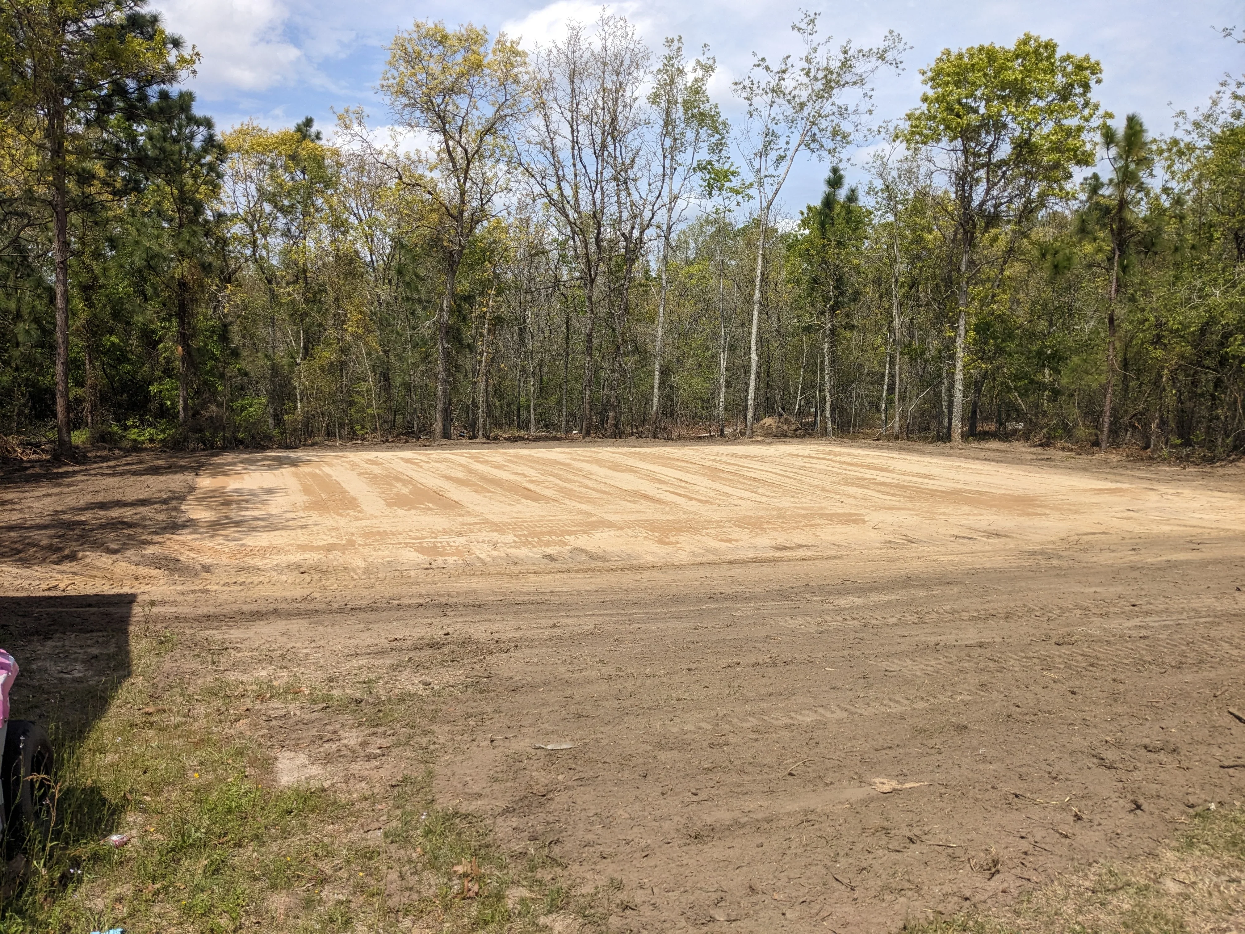 Cleared and graded dirt lot with forest backdrop and visible grading patterns