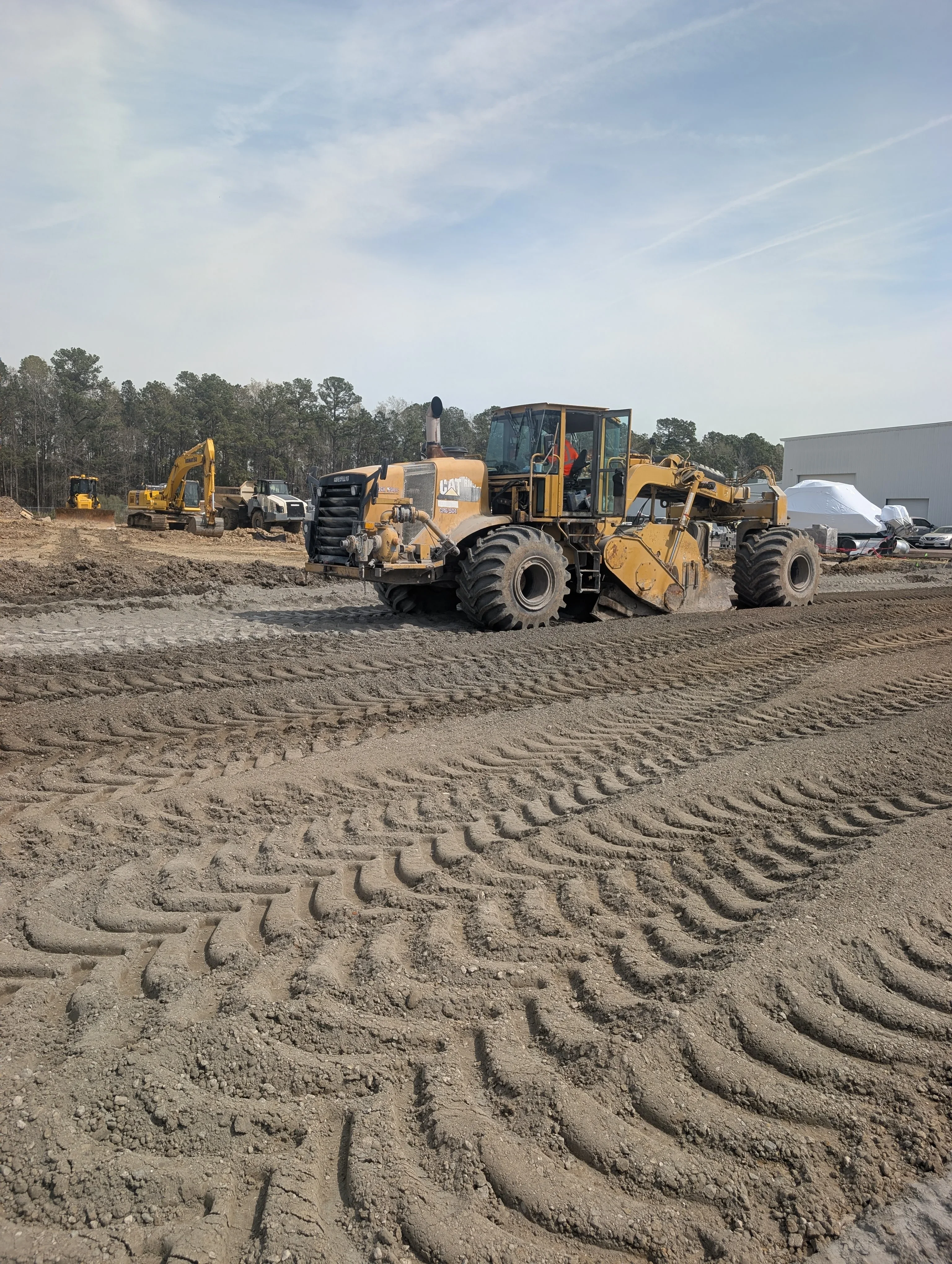 Motor grader creating graded rows in dirt site with heavy equipment and stockpiles