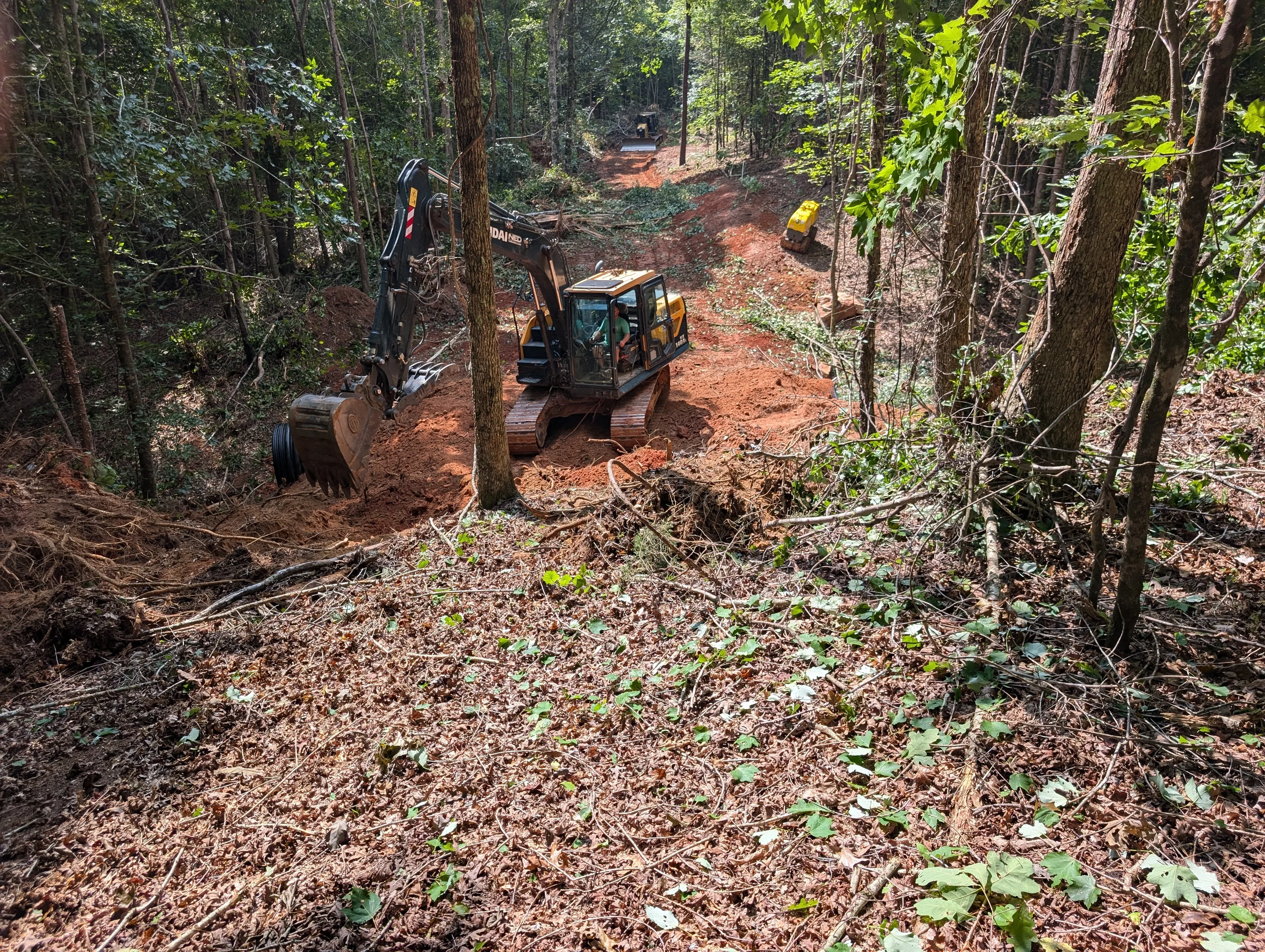 Tracked excavator and mini-excavator grading red clay forest trail through dense timber