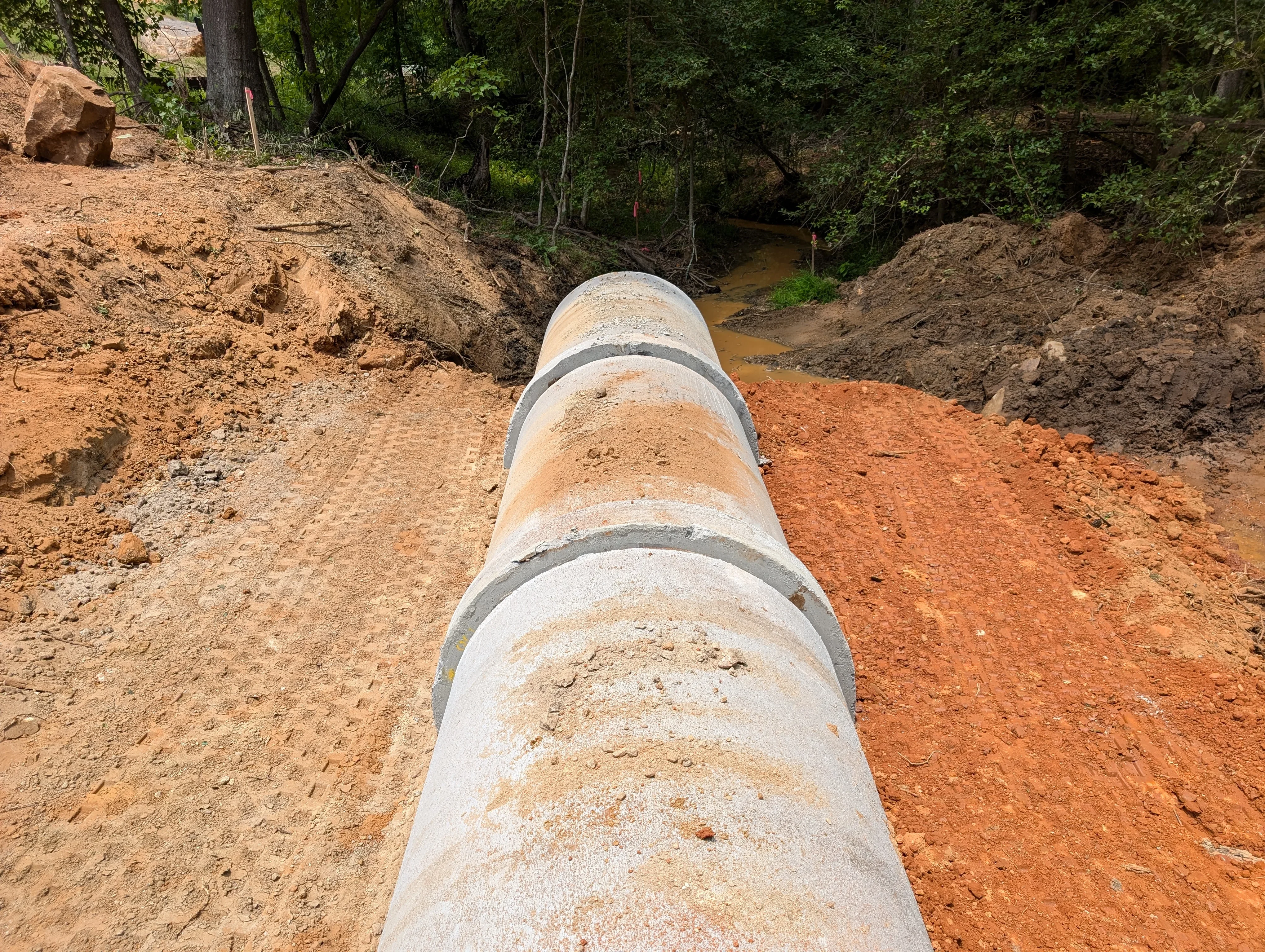 Concrete drainage pipe sections installed in excavated streambed with graded soil and forest backdrop