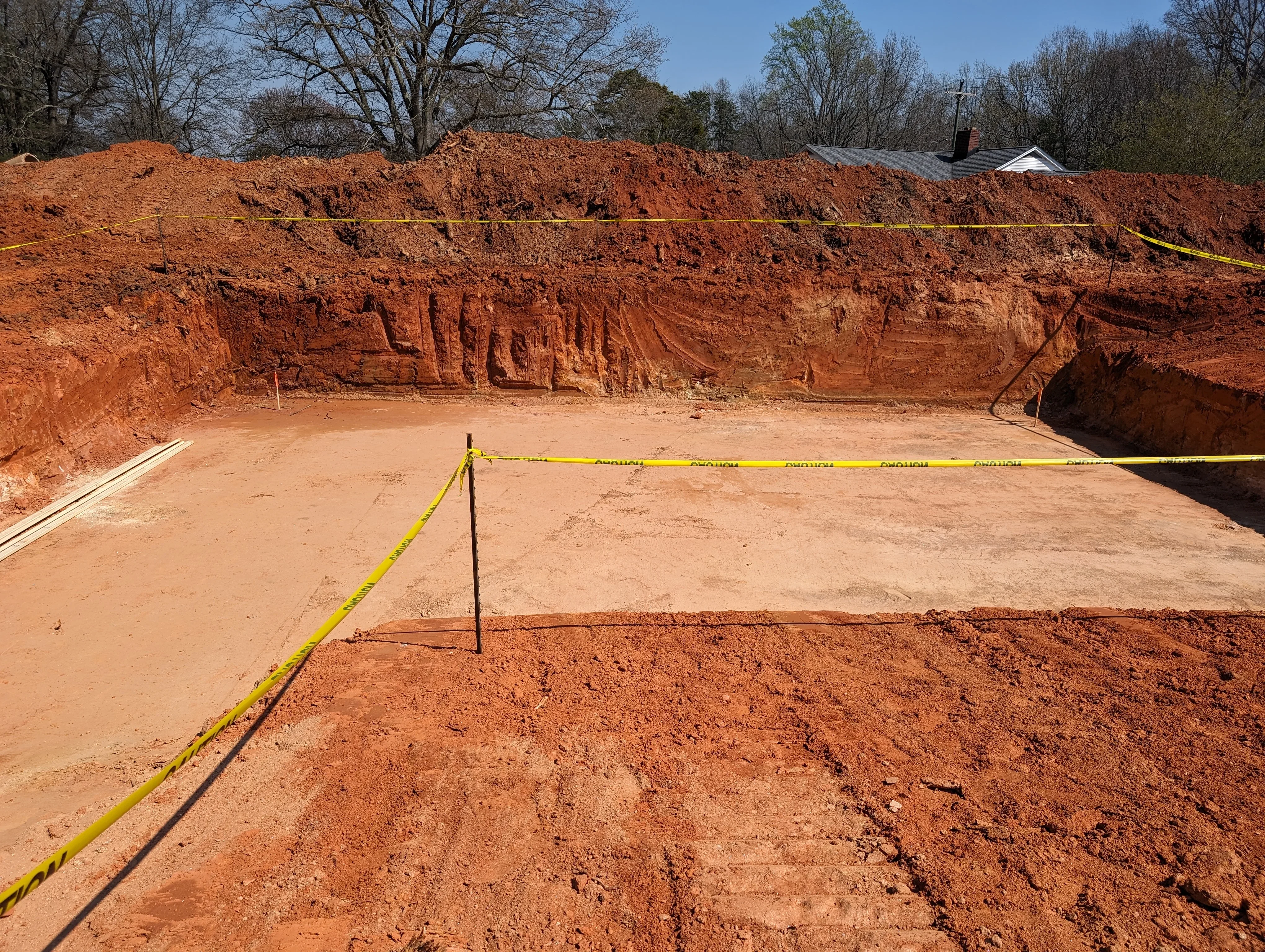 Excavated residential lot with level foundation pad, red clay soil stockpiles, and caution tape