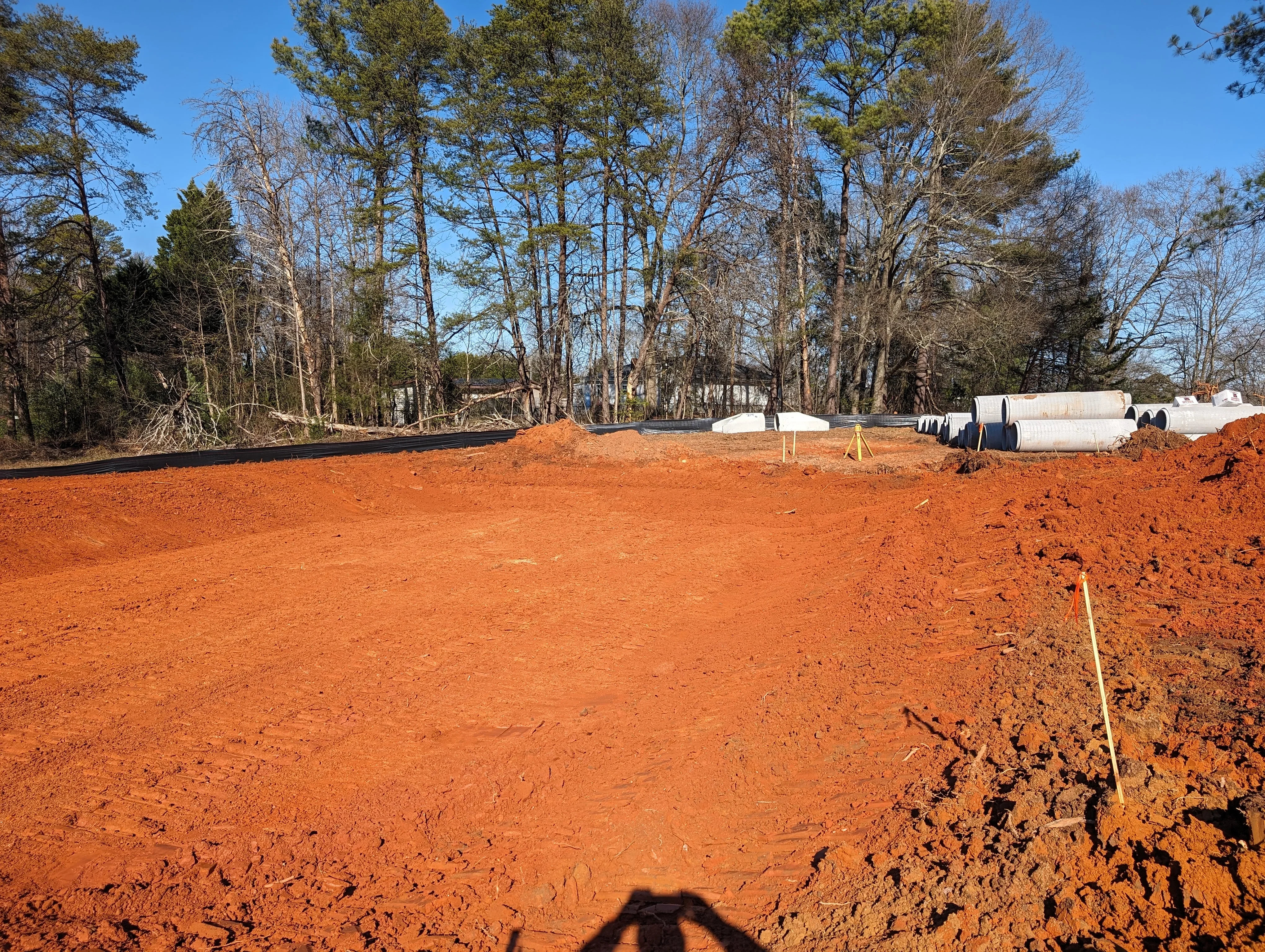 Graded red clay lot with cleared trees, drainage pipes, and construction staging area