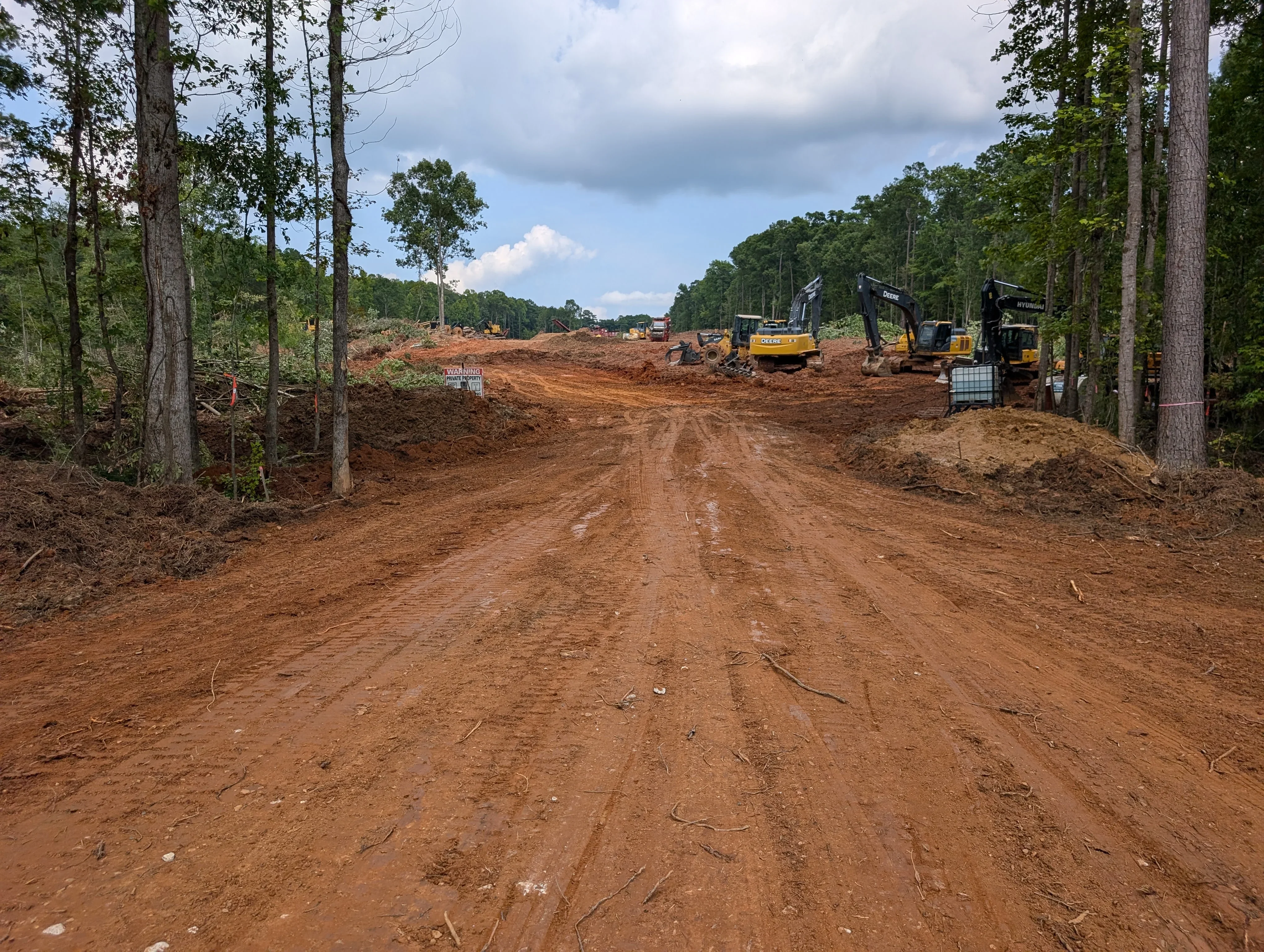 Cleared wooded lot with graded red clay soil, standing trees at edges, and yellow excavators on site