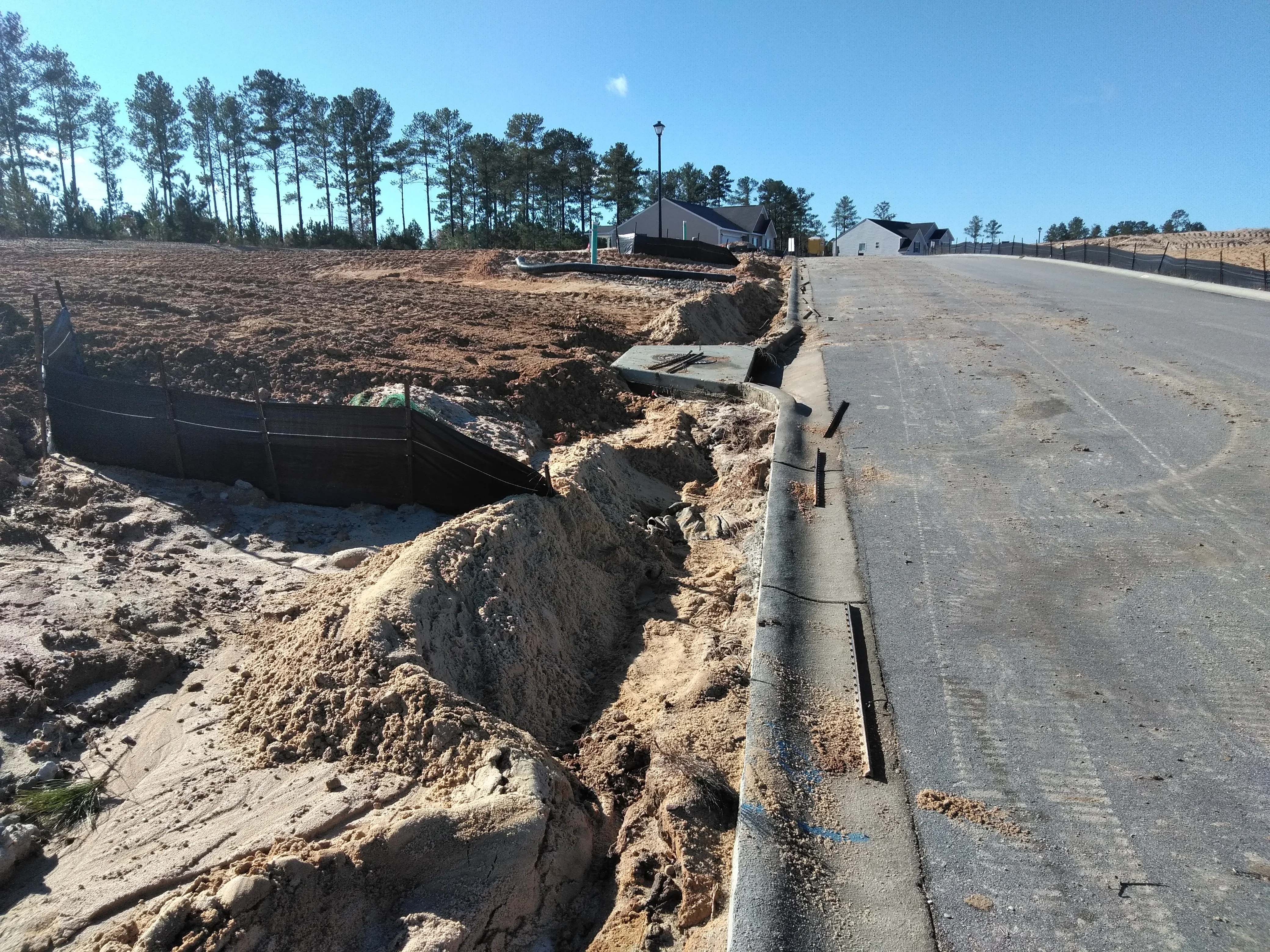 Excavated trench with drainage pipe and erosion control fabric alongside finished concrete driveway