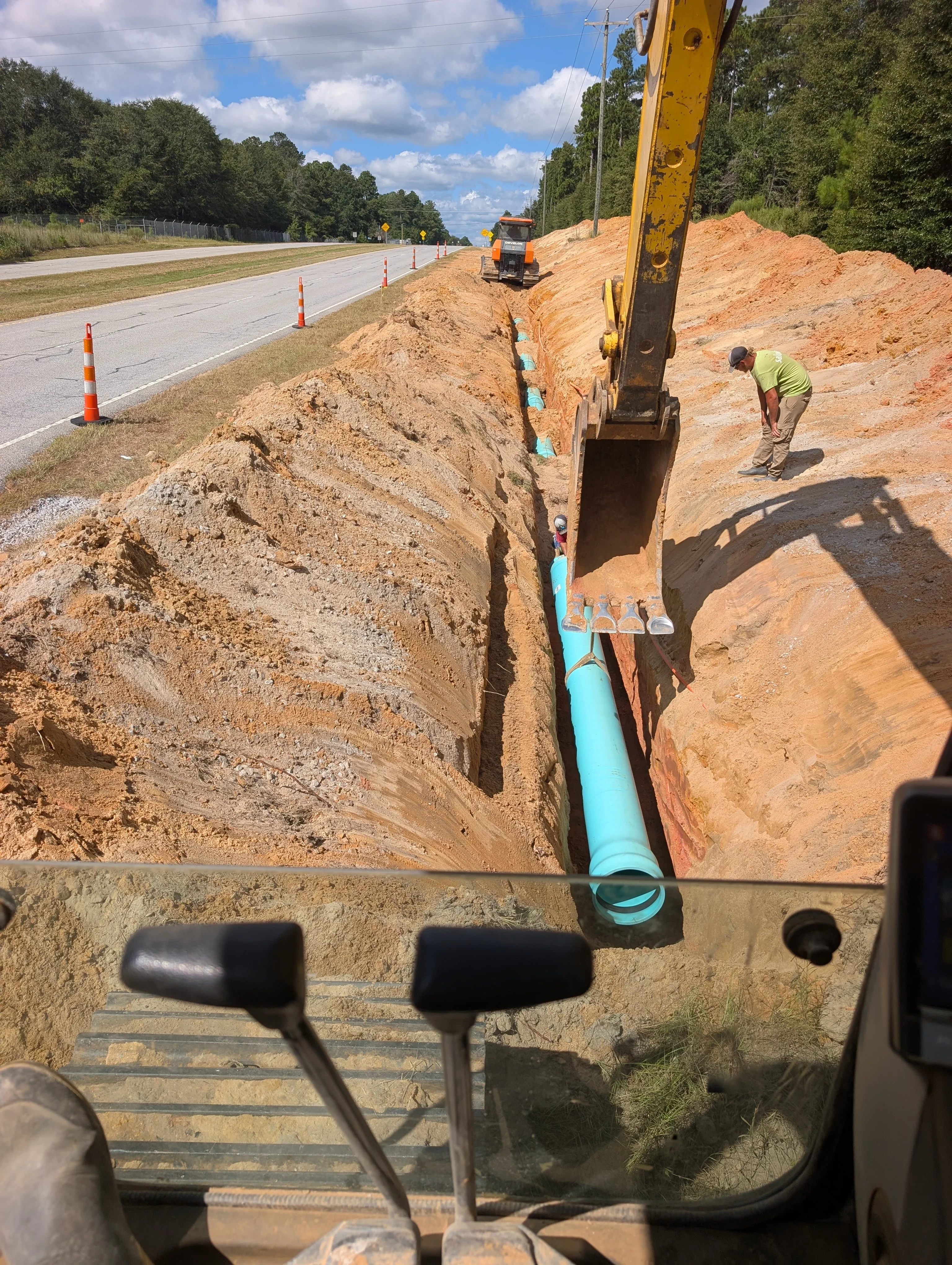 Excavator digging utility trench with blue PVC pipe installation along roadside