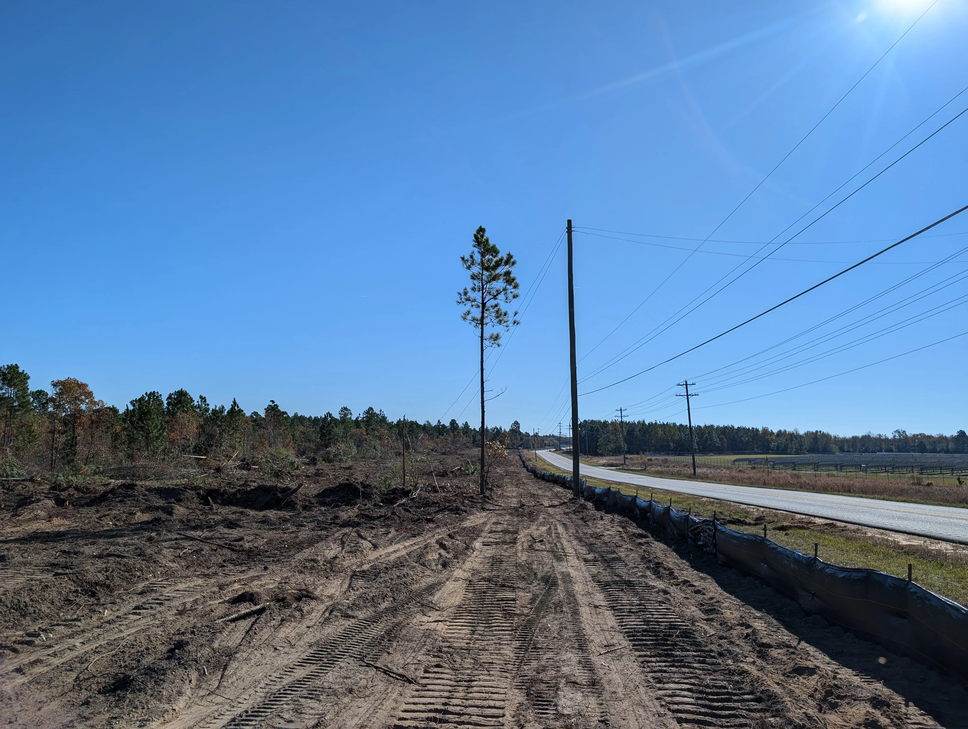 Cleared land with graded dirt, erosion control fencing, and utility poles along paved road